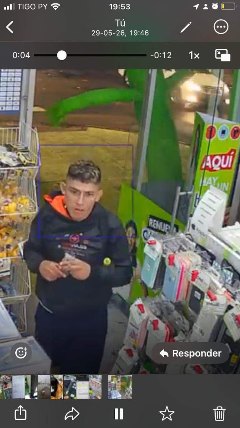 Hombre con sudadera negra y capucha, mirada de sorpresa, en tienda con estanterías de accesorios para móviles.