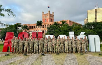Grupo de 30 médicos militares en uniformes posando en formación, ambiente exterior con edificio gubernamental y bandera paraguaya.