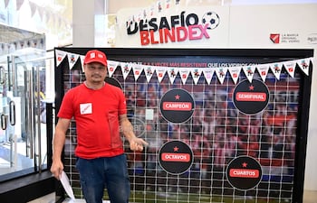Hombre de mediana edad en camiseta y gorra rojas, frente a decoraciones del evento Desafío Blindex, sosteniendo un papel.