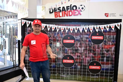 Hombre de mediana edad en camiseta y gorra rojas, frente a decoraciones del evento Desafío Blindex, sosteniendo un papel.