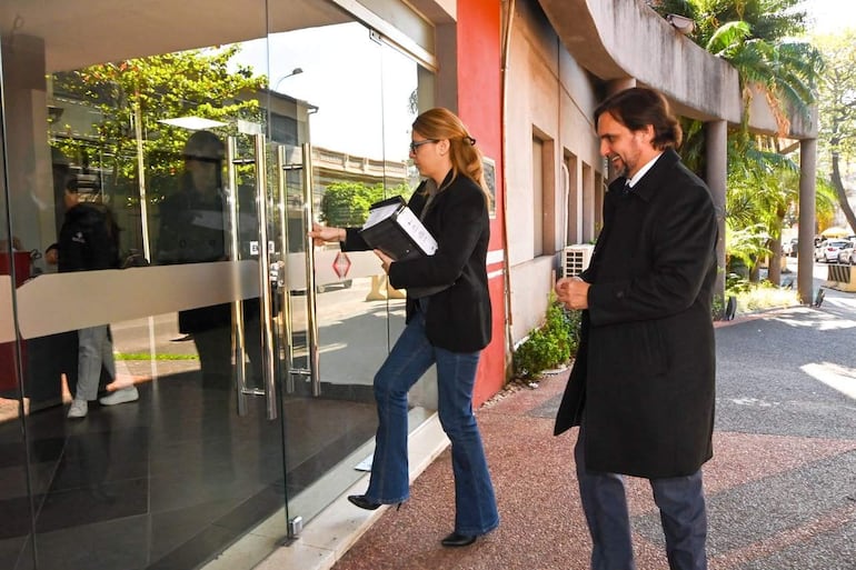 Melissa Ramírez sostiene documentos frente a una puerta, vestida con chaqueta y jeans; Jorge Sabaté la observa sonriente con abrigo oscuro.
