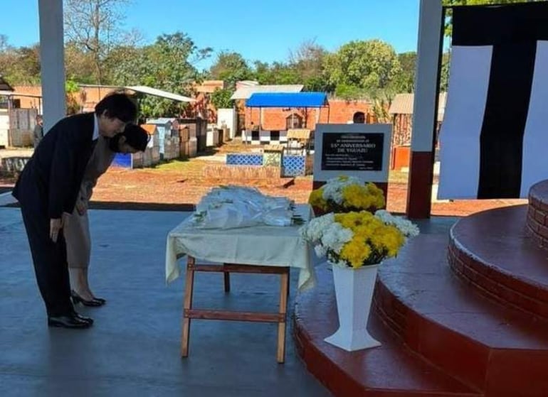 Un hombre en traje oscuro y una mujer formal se inclinan en un ambiente con flores amarillas, rodeados de lápidas.