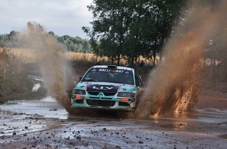 Un Mitsubishi en movimiento, salpicando barro y agua, rodeado de vegetación en una pista de rally.