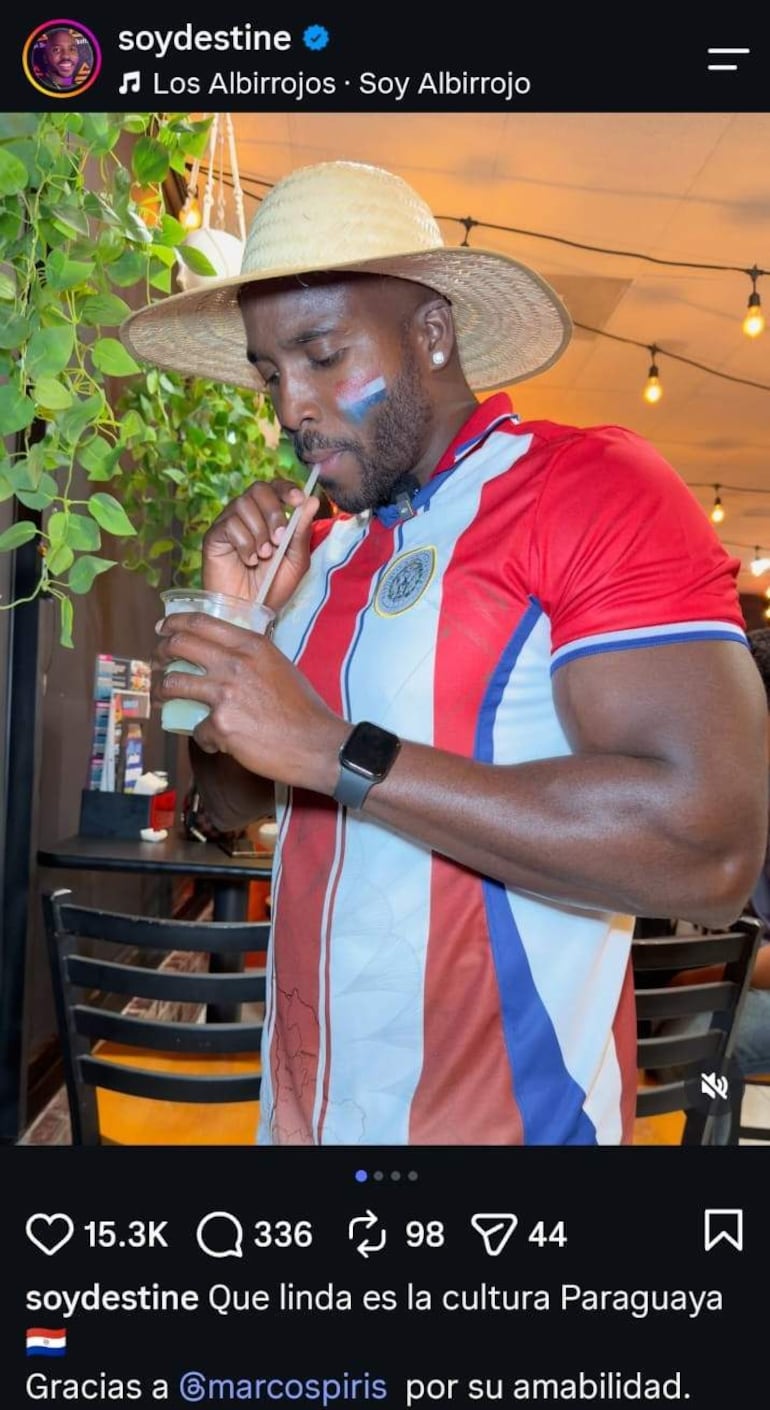 Yonathan Destine viste camiseta de Los Albirrojos y sombrero de paja, sosteniendo una bebida, con expresión feliz en un café.