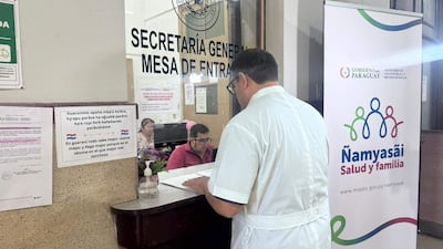 Hombre de espaldas con bata blanca gestiona documentos, mientras mujer observa detrás del mostrador en ambiente administrativo colorido.