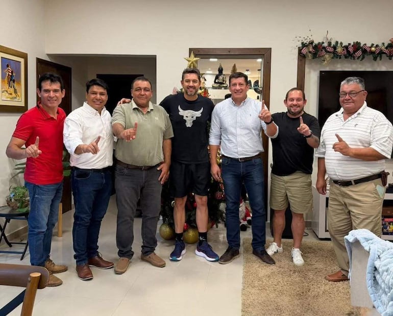 Alliana con remera negra y Arce en camisa celeste, todos sonrientes y levantando un dedo en un ambiente decorado navideño.