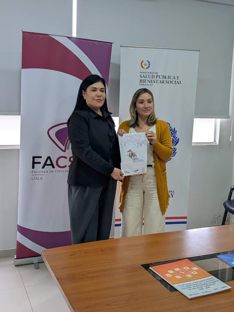 Dos mujeres, una en abrigo beige y la otra en camisa oscura, sostienen un documento en una oficina con banners del MSP y FACSO.