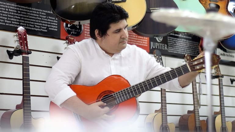 Jorge Montefilpo en camisa blanca, concentrado, toca guitarra roja en una tienda con otras guitarras colgadas.
