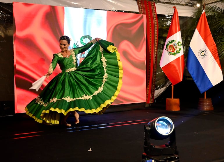 Mujer con vestido verde adornado baila, sosteniendo un pañuelo, con banderas de Perú y Paraguay de fondo en un ambiente festivo.