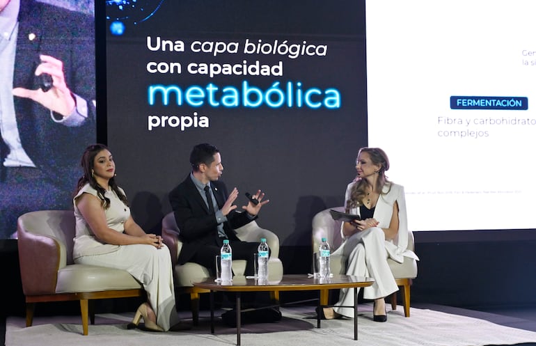 Carolina Sosky en vestido blanco observa a un hombre con traje negro que presenta, mientras otra mujer anota en una tablet.