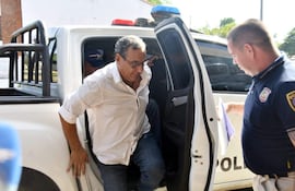 Un hombre con camisa blanca y jeans desciende de un vehículo policial, mientras un oficial uniformado revisa documentos a su lado.