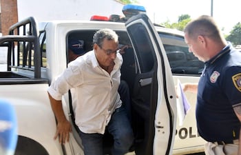 Un hombre con camisa blanca y jeans desciende de un vehículo policial, mientras un oficial uniformado revisa documentos a su lado.