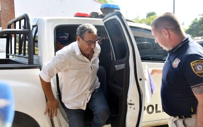 Un hombre con camisa blanca y jeans desciende de un vehículo policial, mientras un oficial uniformado revisa documentos a su lado.