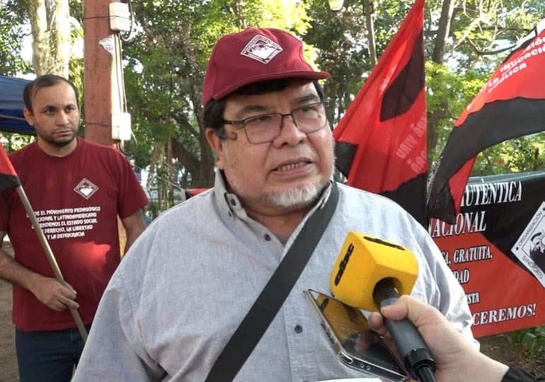 Gabriel Espínola frente a micrófono amarillo y gorra roja habla enérgicamente, mientras un hombre con camiseta roja sostiene una bandera detrás.