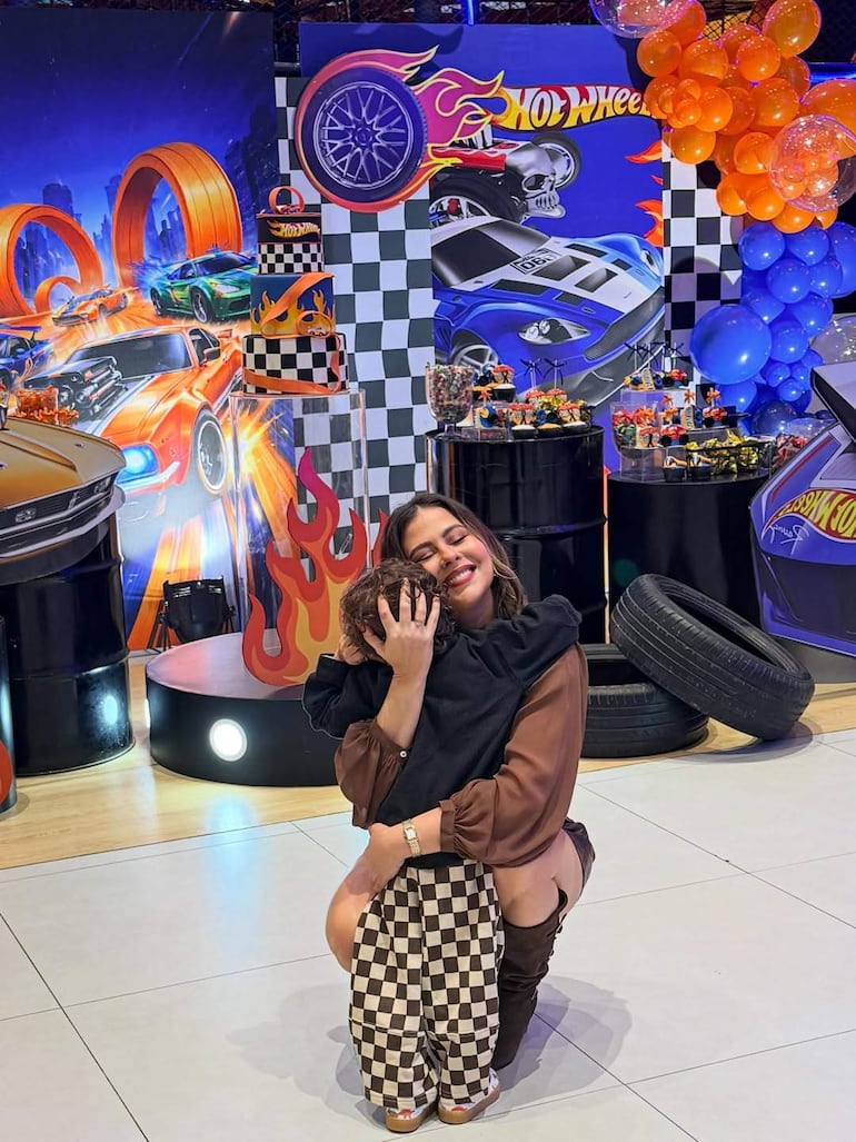 Mujer de cabello largo abrazando a un niño en fiesta temática de Hot Wheels, rodeados de globos naranjas y azules.
