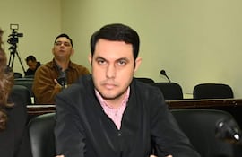 Hernán David Rivas con chaqueta negra y camisa de rayas, sentado junto a una mujer con blusa oscura en sala de reunión.