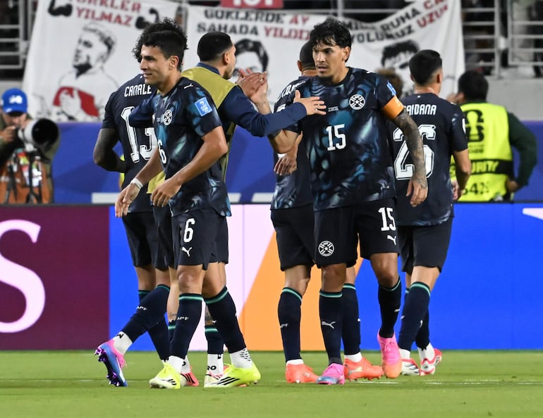Jugadores de Paraguay en camisetas negras con escudo, uno recibe una palmada en el hombro mientras celebran juntos en el campo.