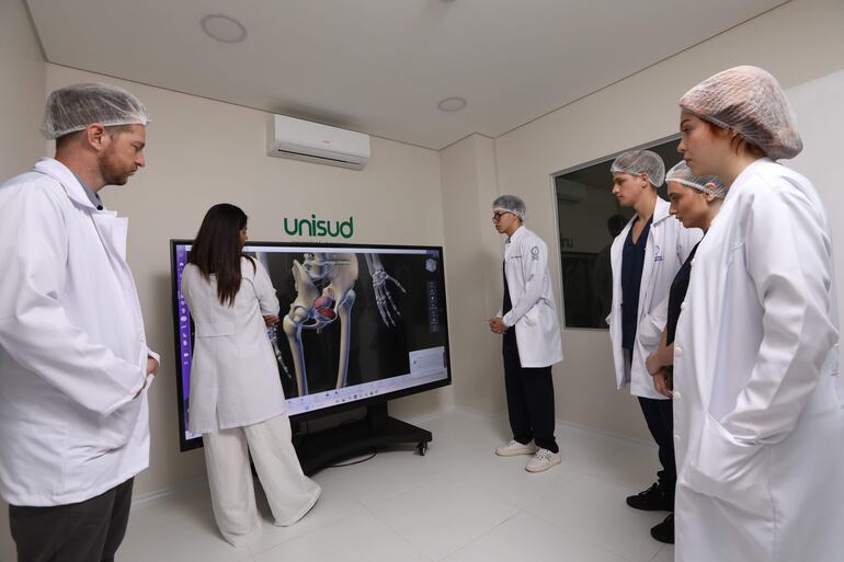 Dos mujeres y tres hombres en aulas claras con batas blancas, una mujer presenta en un monitor sobre anatomía humana.