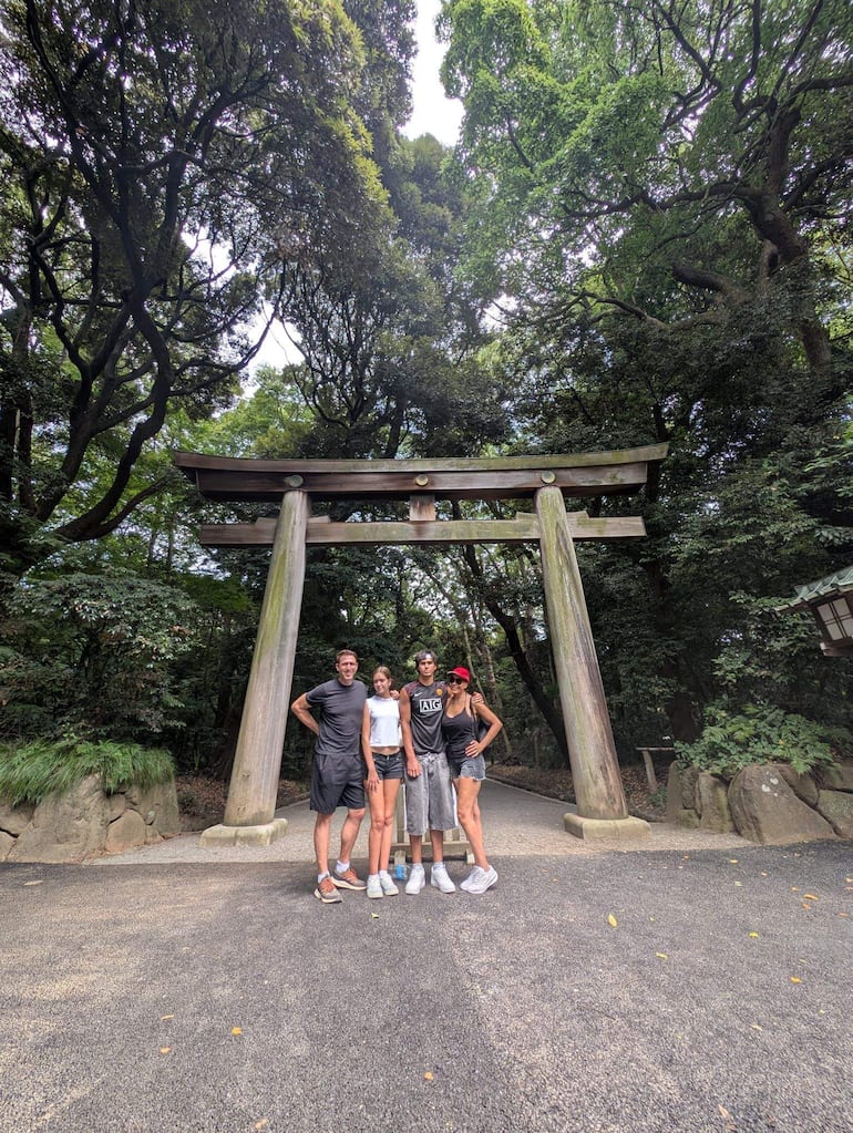 Cuatro individuos sonrientes, dos hombres y dos mujeres, frente a un torii verde en un bosque denso.