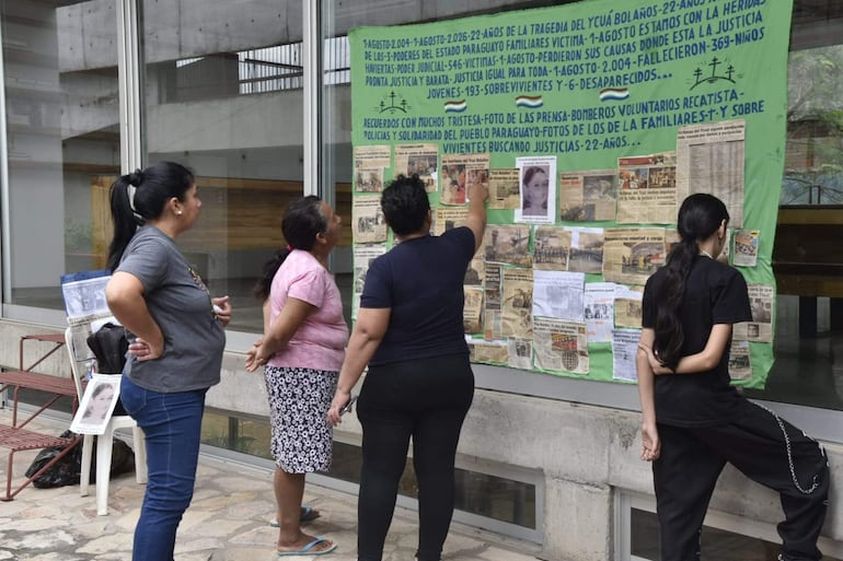 Cuatro mujeres, una en camiseta gris, otra en rosa, y otra en negra, observan un panel con fotos y textos conmemorativos.