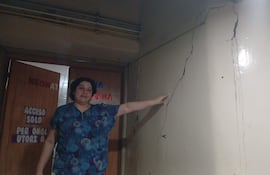 Mujer con blusa azul señala grieta en la pared, en un pasillo con letreros de acceso restringido.