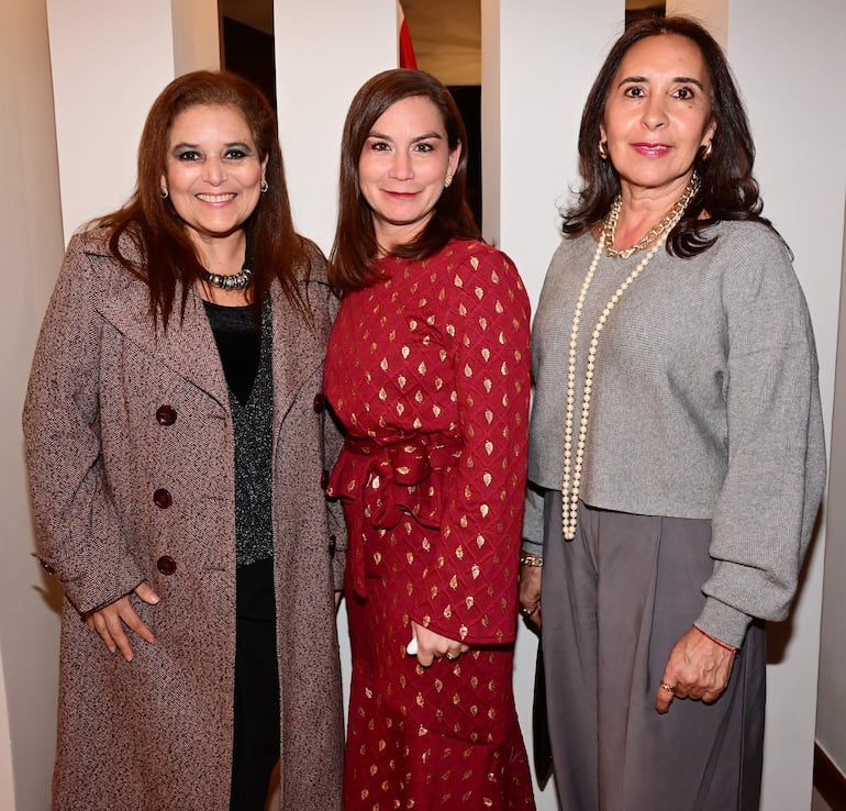 Tres mujeres sonrientes: una con abrigo marrón, otra en vestido rojo y la última en suéter gris, en un entorno elegante.