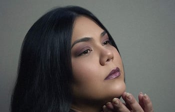 Mujer de cabello largo y oscuro con expresión pensativa, manos cerca del rostro en un fondo neutro.