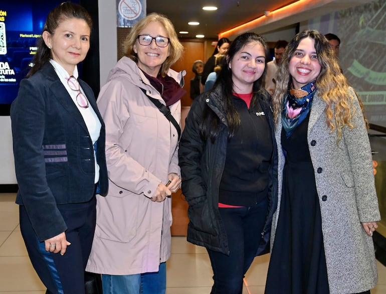Cuatro mujeres conversan en un auditorio; una con saco oscuro, otra con abrigo beige, sonriendo y transmitiendo alegría.