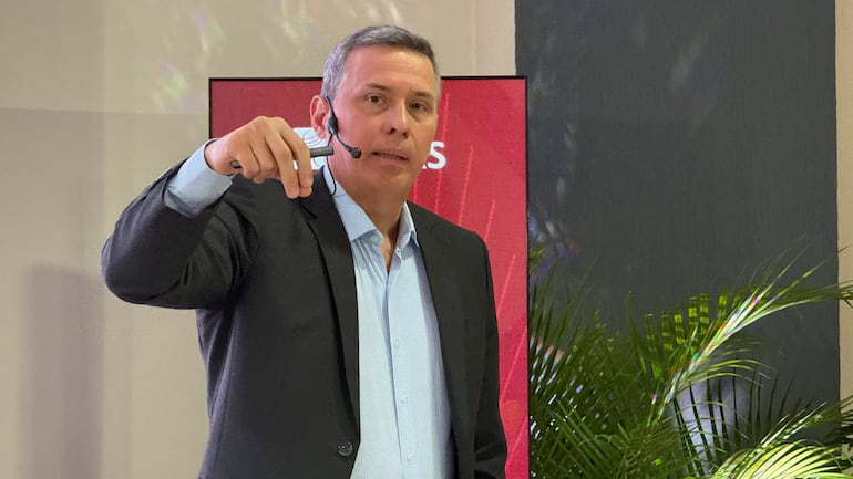 Hombre canoso en traje oscuro y camisa clara, sostiene un objeto y habla ante un panel rojo decorado con plantas.