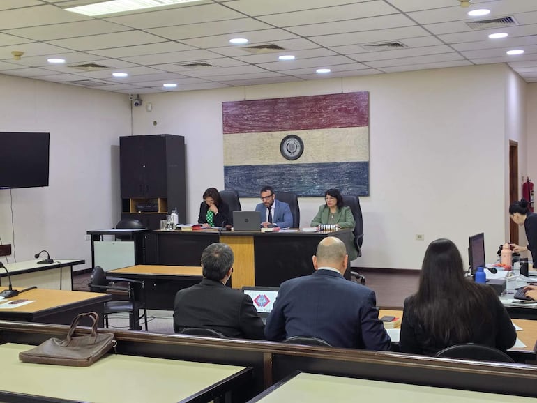 Tres personas en escritorio, con una mujer de blusa negra, hombre con traje oscuro y mujer en chaqueta verde, en una sala con decoración paraguaya.
