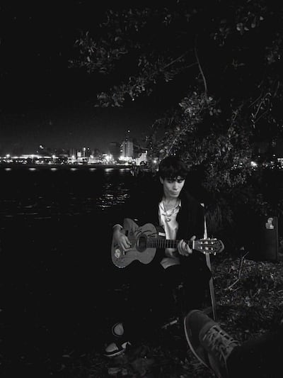 Lucas Montanaro, vestido con camiseta blanca y chaqueta oscura, toca la guitarra en un ambiente iluminado y tranquilo, con un espectador visible.