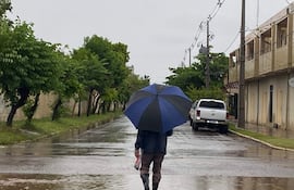 Persona con abrigo oscuro y botas de goma sostiene paraguas negro y azul en calle inundada rodeada de árboles y edificios.