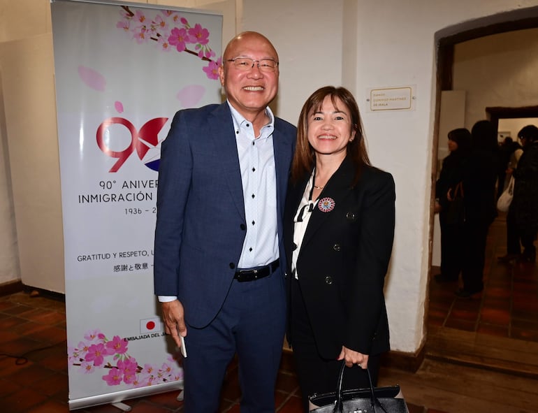 Hombre calvo en traje oscuro y mujer con chaqueta oscura sonríen en un evento de la embajada japonesa, rodeados de un ambiente formal.