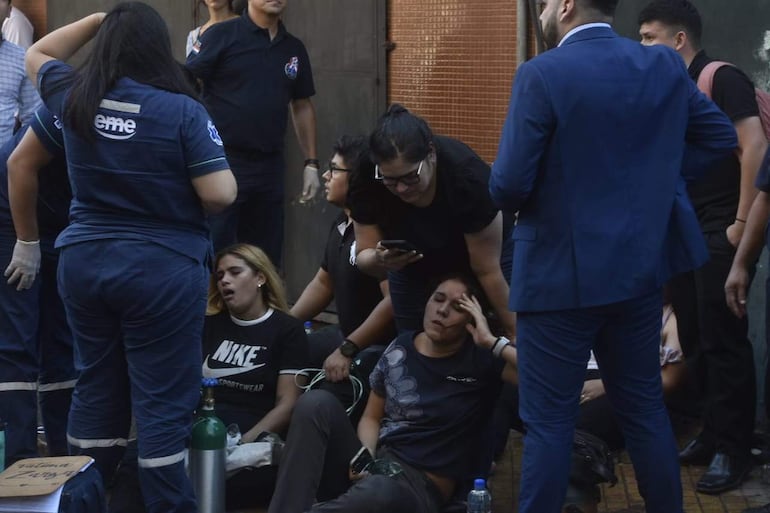 Dos mujeres sentadas en el suelo, una con camiseta Nike, reciben asistencia médica mientras un hombre observa. Cilindro de oxígeno presente.