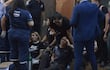 Dos mujeres sentadas en el suelo, una con camiseta Nike, reciben asistencia médica mientras un hombre observa. Cilindro de oxígeno presente.