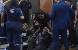 Dos mujeres sentadas en el suelo, una con camiseta Nike, reciben asistencia médica mientras un hombre observa. Cilindro de oxígeno presente.