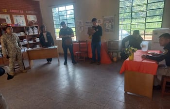 Hombre de uniforme militar camuflado y cuatro personas con camisetas negras conversan en ambiente interior con mesa y flores.