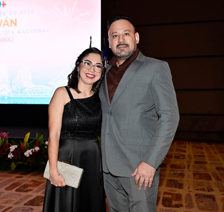 Mujer con vestido negro brillante y hombre en traje gris sonríen en evento bien iluminado con fondo festivo.