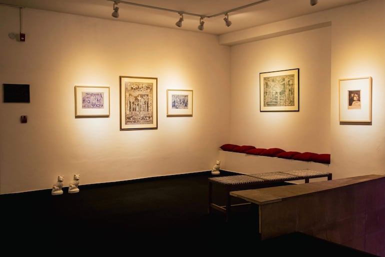 Espacio de galería con obras decorativas en las paredes, un banco rojo y esculturas de cerámica en una iluminación cálida.