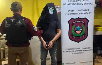 Funcionario de seguridad en chaleco negro sujeta a un hombre con saco negro y brazos esposados en una cocina.