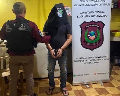 Funcionario de seguridad en chaleco negro sujeta a un hombre con saco negro y brazos esposados en una cocina.
