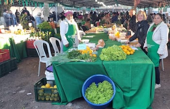 Dos vendedoras con delantales verdes sonríen detrás de una mesa de vegetales en un mercado comunitario, con gente comprando de fondo.