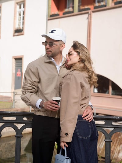 Pareja sonriente junto al canal, el hombre con chaqueta beige y gorro blanco, la mujer con chaqueta beige y jeans oscuros.