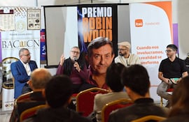 Panel de discusión con cuatro hombres: orador central con gafas, bufanda oscura y manos en movimiento.