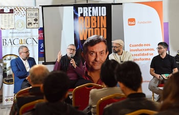 Panel de discusión con cuatro hombres: orador central con gafas, bufanda oscura y manos en movimiento.