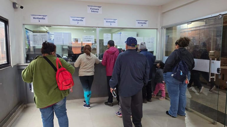 Mujeres y niños en fila frente a ventanillas de farmacia, con expresiones de preocupación y impatiencia.