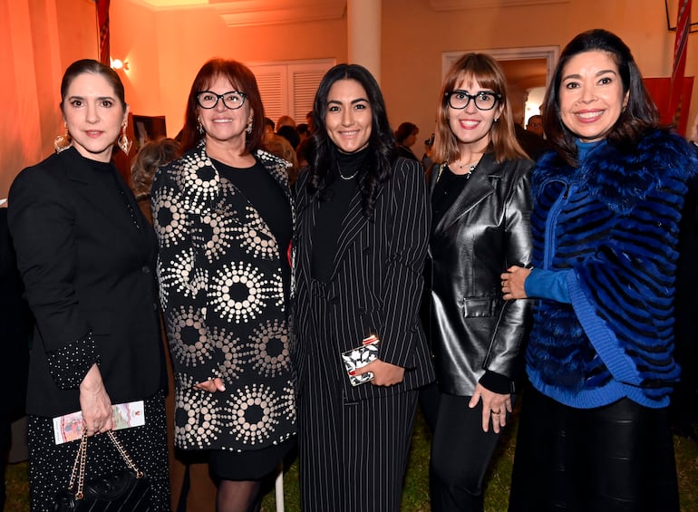 Cinco mujeres bien vestidas, con abrigos y chaquetas, posan sonrientes en un ambiente festivo con decoración cálida.
