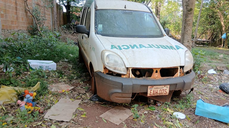 Ambulancia deteriorada en un terreno cubierto de hierba, rodeada de escombros y cajas, sin personas visibles.