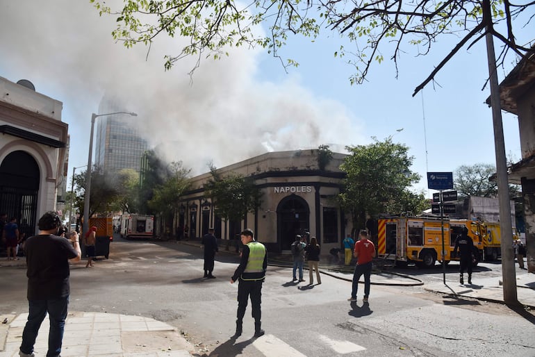 Hombre con camiseta verde de seguridad supervisa incendio en local Napoles, mientras bomberos trabajan en el fondo y humo se eleva.
