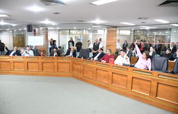 Seis personas sentadas alrededor de una mesa en forma de 'U', algunas levantando la mano. Mujer en blusa rosa parece querer participar.